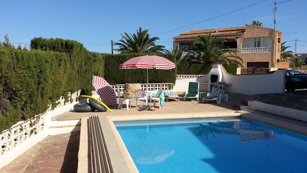 A swimming pool with lounge chairs and umbrellas, a stone wall, and a building with a tiled roof in the background. - 5 DBR villa.Dual level with own pool. Walking distance to Calpe's Levante beach (Alicante)