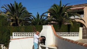 A man using a barbecue grill on a patio with a white wall, palm trees, and a building in the background. - 5 DBR villa.Dual level with own pool. Walking distance to Calpe's Levante beach (Alicante)