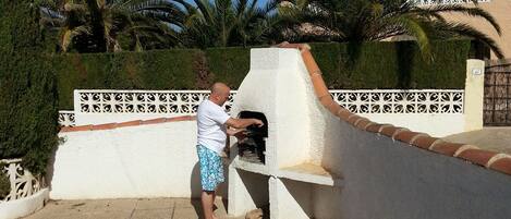 A man using a barbecue grill on a patio with a white wall, palm trees, and a building in the background.