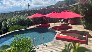 Un espace piscine agrémenté de parasols rouges, d’une terrasse en bois et d’un mur en pierre.