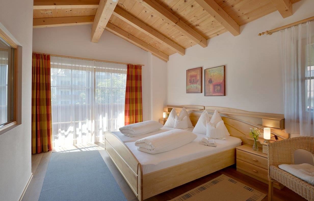 A hotel room with a wooden ceiling, a large bed with white linens, a bedside table with a lamp, and a wicker chair.