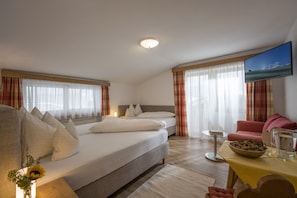A hotel room with a large bed, a sofa, a small table with a bowl of fruit, a pitcher of water, and a television on the wall. - Hotel Theresia Garni (Sankt Johann in Tirol)