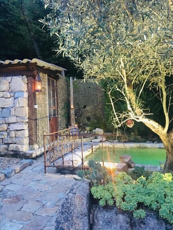 A stone building with a metal fence, a small pond, and a tree with hanging ornaments.