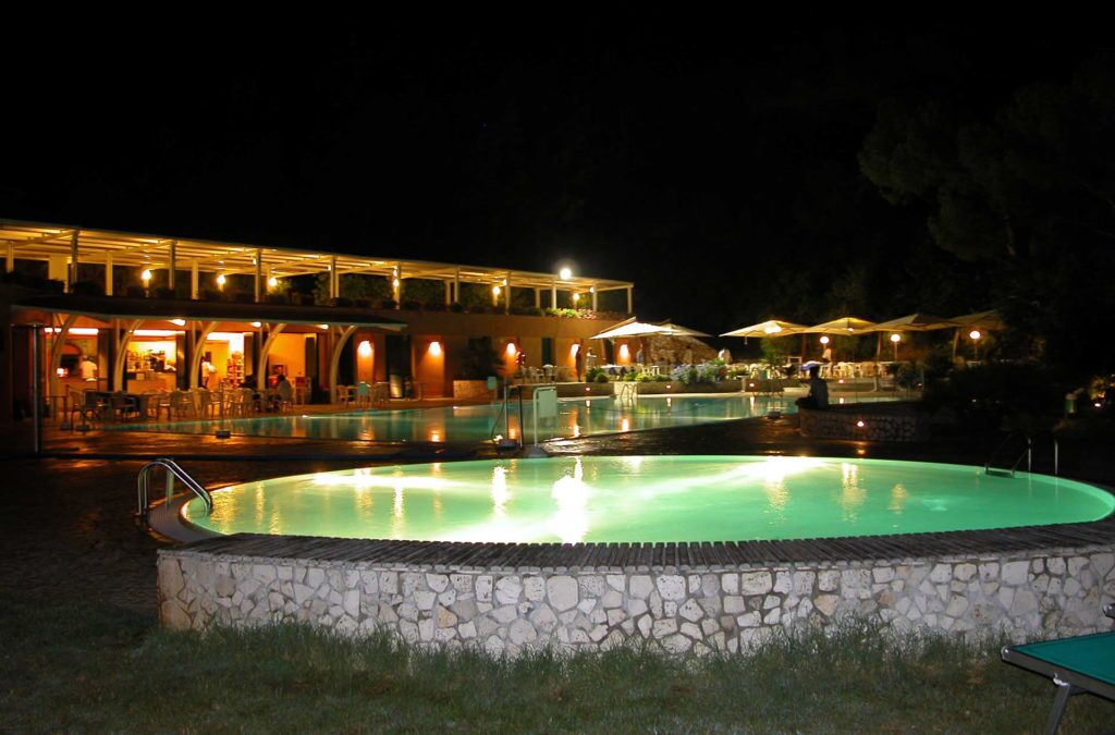 A nighttime pool area with a stone border, a building with multiple levels and outdoor seating, and a well-lit pool.