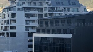 Un edifici de diverses plantes amb balcons i grans finestres, amb un turó i un cel amb núvols com a fons.