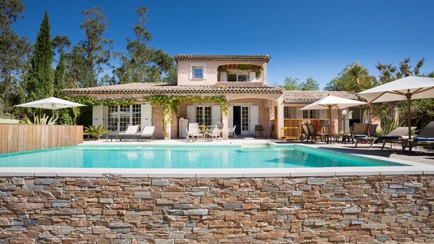 Une maison à deux étages avec une piscine, du mobilier d’extérieur et un mur en pierre.
