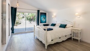 Une chambre avec un grand lit, une table de chevet, une lampe fixée au mur et une vue sur un jardin, accessible par une porte-fenêtre coulissante.