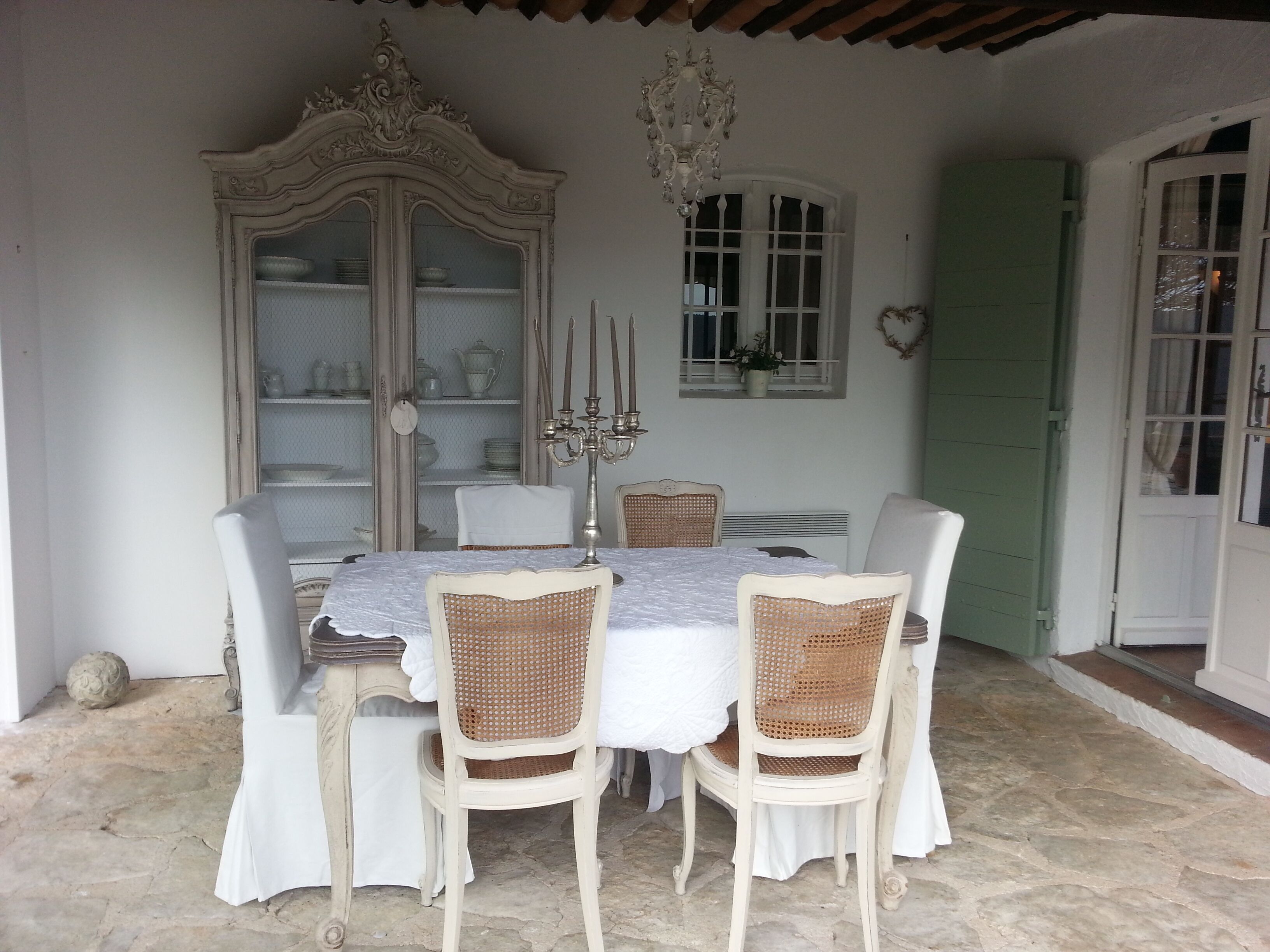 A dining area with a table set for four, surrounded by wicker chairs, a white cabinet with glass doors, and a chandelier.