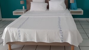 A neatly made bed with white linens and a decorative border, a wooden headboard, and a teal-colored wall with two decorative mirrors.