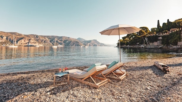 A beach with lounge chairs and an umbrella, rocky shoreline, boats, and hills in the background.