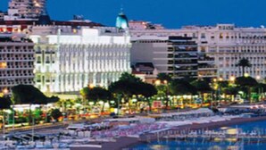 A coastal cityscape with a beachfront promenade, buildings, and a large cruise ship docked nearby.