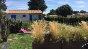 Zona residencial con una casa blanca con contraventanas azules, techo rojo y jardín con diversas plantas y una silla rosa.