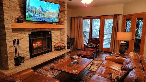 A cozy living room with a fireplace, leather sofa, and a television mounted on a brick wall. - Ski In/Ski Out + Private Hot Tub + Sleeps 8 + Free Parking  (Keystone)