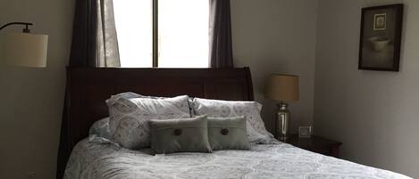 A bedroom with a large bed, a window with curtains, a bedside lamp, and a framed picture on the wall.