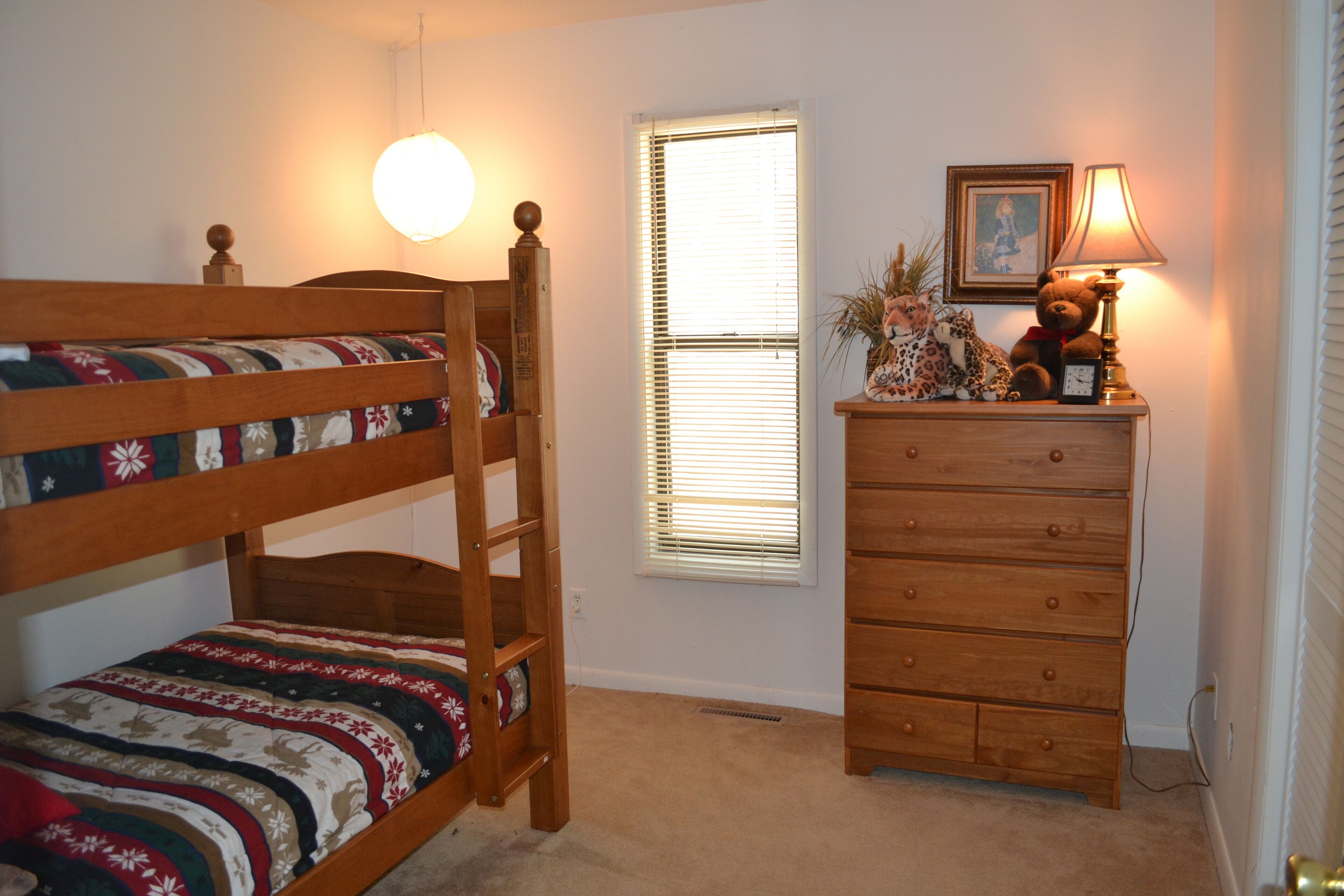 A bedroom with a bunk bed, a dresser, a lamp, and a window with blinds.