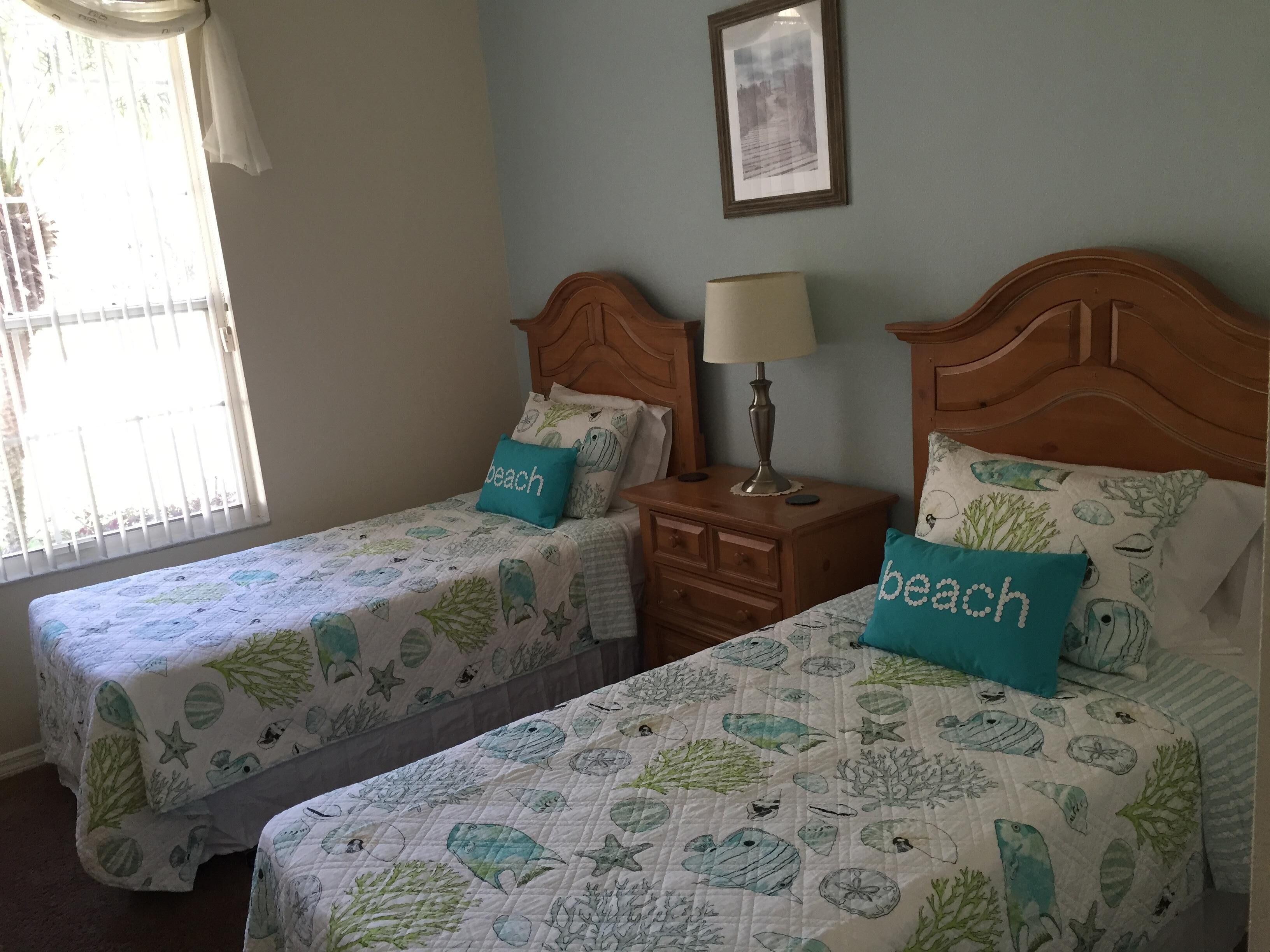 A bedroom with two beds, each with a beach-themed bedspread and matching pillows. There is a nightstand with a lamp between the beds, and a framed picture on the wall above the nightstand.