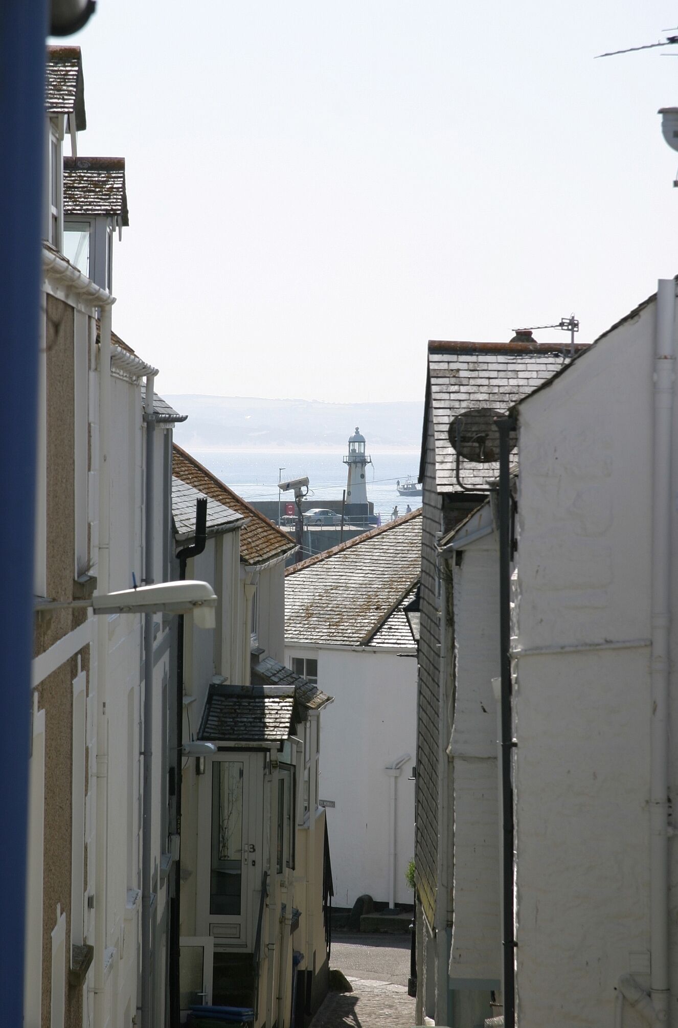 Eine schmale Straße mit Gebäuden an beiden Seiten, ein Leuchtturm in der Ferne und freie Sicht auf das Meer.