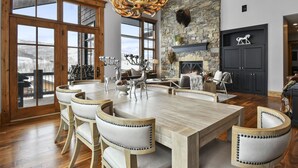 Amplia sala de estar con una gran mesa de madera para comer, sillas de cuero, una chimenea de piedra y vistas a montañas nevadas a través de grandes ventanales.