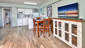 Una cocina moderna con electrodomésticos blancos, una zona de comedor con sillas de madera y un televisor sobre un mueble.