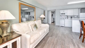Un salón moderno con un sofá blanco, dos lámparas y un espejo grande. Se ve una cocina con electrodomésticos blancos al fondo.