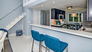 A modern kitchen with a white countertop, black cabinets, and stainless steel appliances. There are two blue bar stools and a white chair. - Home Away From Home! 3 Bedroom Home Completely Updated; community pool access and BHI club access (Bald Head Island)