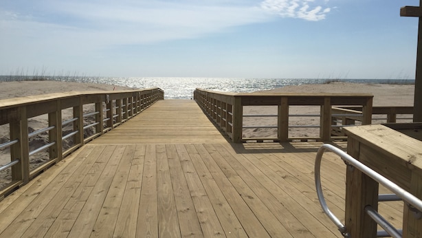 Een houten looppad dat naar een strand leidt, met metalen leuningen aan beide zijden.