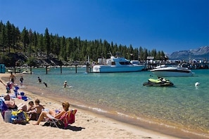 En strand med kristallklart turkost vatten, en brygga med båtar och människor som kopplar av på sanden.