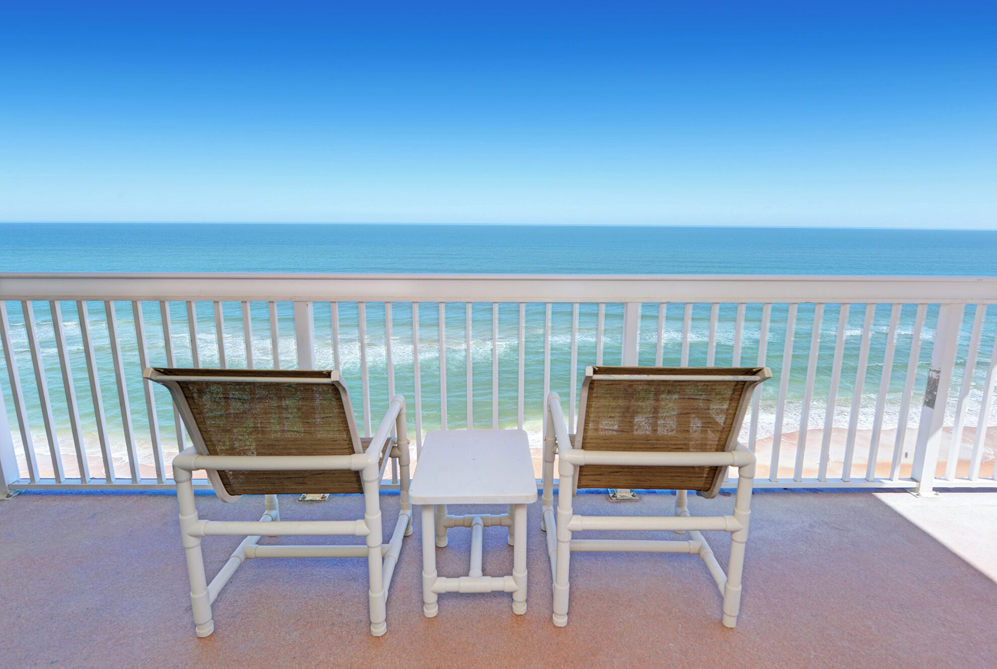 Twee witte rieten stoelen met een klein witte tafeltje op een balkon met een witte reling, met uitzicht op een strand en de oceaan.