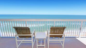 Twee witte rieten stoelen met een klein witte tafeltje op een balkon met een witte reling, met uitzicht op een strand en de oceaan.