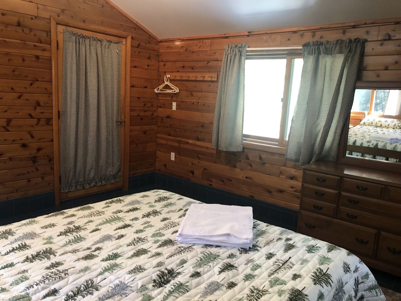 A wooden cabin bedroom with a bed, a dresser, and a window with curtains.