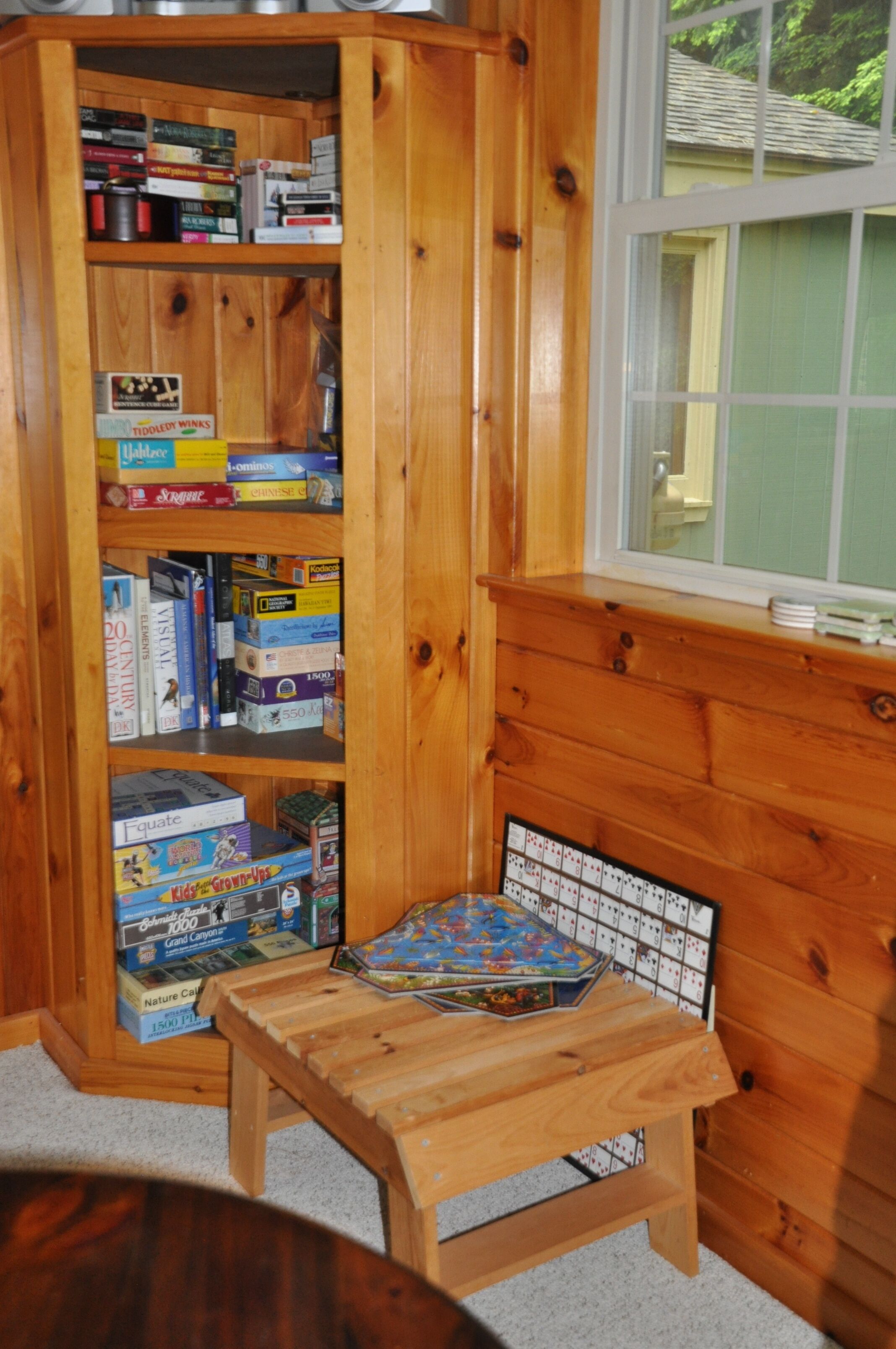 Una estantería de madera llena de juegos de mesa y una pequeña mesa de madera con un juego de mesa frente a una ventana.