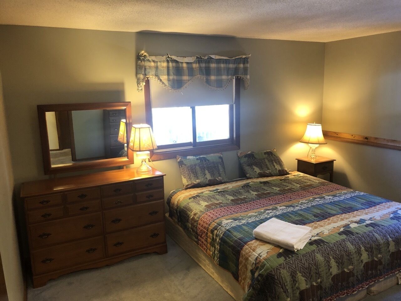 A bedroom with a bed, dresser, two bedside lamps, a window with curtains, and a mirror.
