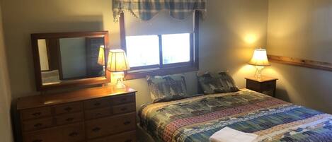 A bedroom with a bed, dresser, two bedside lamps, a window with curtains, and a mirror.