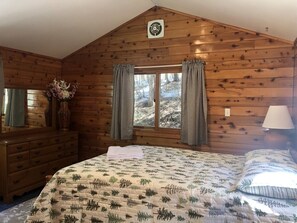 Une chambre dans une cabane en bois, avec un lit, une commode, une fenêtre avec des rideaux et une lampe.