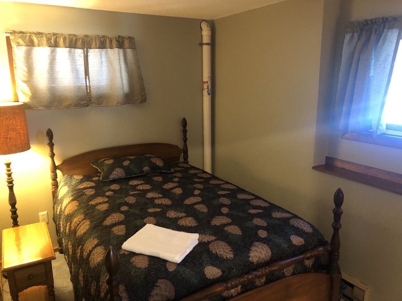 A bedroom with a bed, a nightstand, a lamp, a window with curtains, and a wall-mounted pipe.