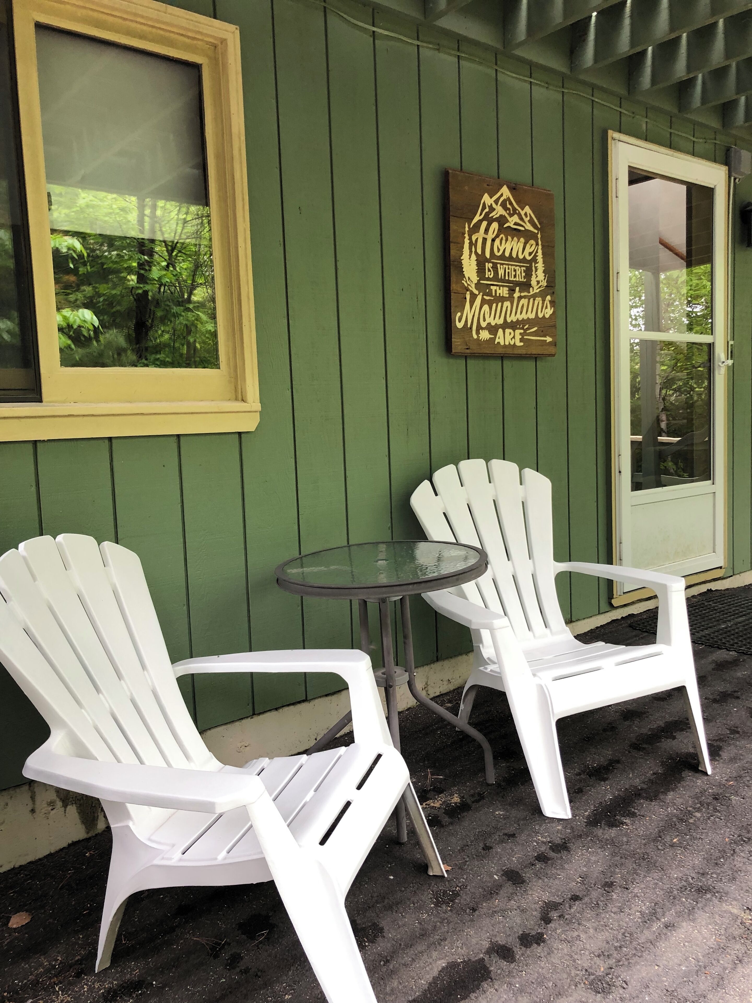 Two white Adirondack chairs and a glass table outside a building with green walls and a wooden sign that reads "Home is where the mountains are."