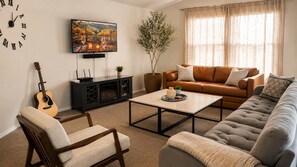 A living room with a sofa, armchair, coffee table, TV, guitar, and a plant.