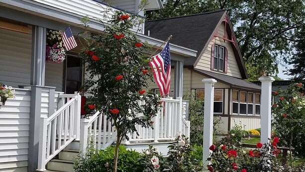 Ein zweistöckiges Haus mit Veranda, einem kleinen Garten mit roten Blüten und einer amerikanischen Flagge.