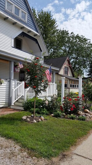 A two-story house with a porch, a small garden with red flowers, and an American flag. - 1 BLOCK TO TOWN ~ 5 BLOCKS TO BEACH! Leave your car at home for the duration! (South Haven)