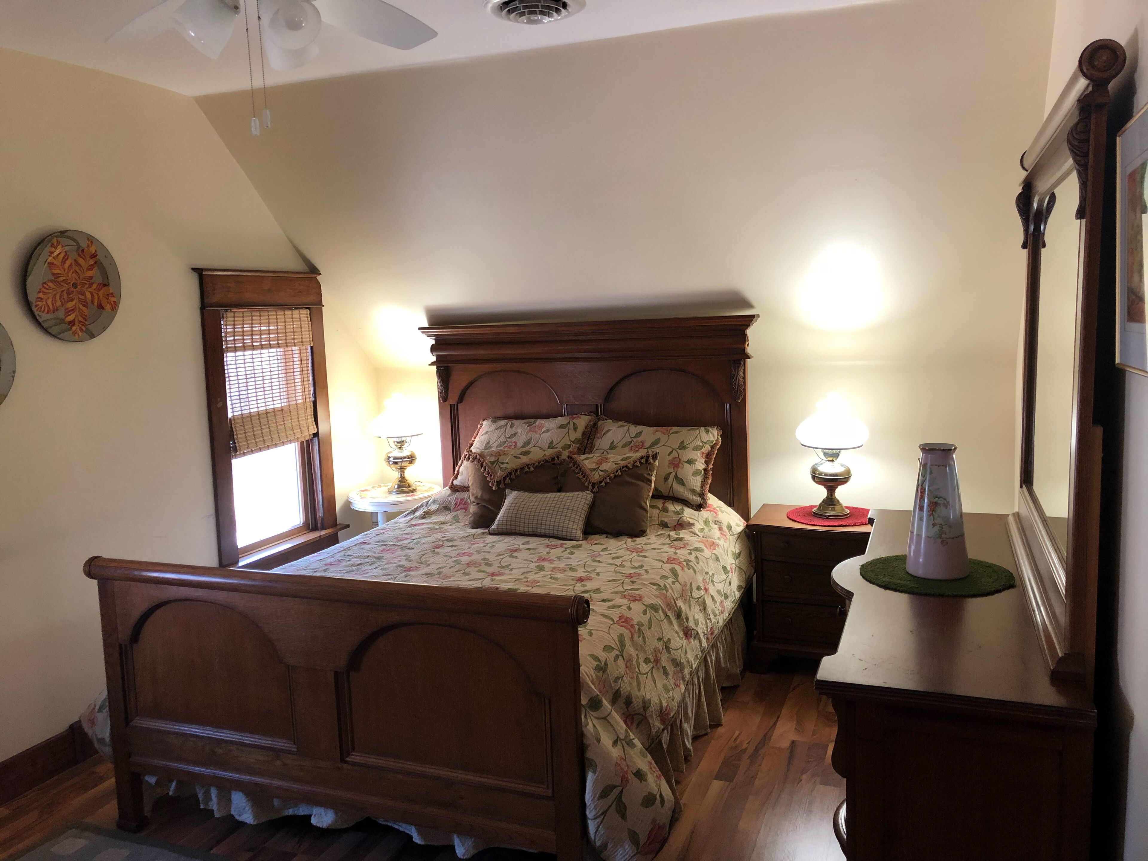 A bedroom with a wooden bed, a nightstand with a lamp, a mirror, and a window with blinds.