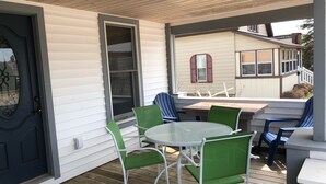 Eine überdachte Terrasse mit einem runden Tisch und grünen Stühlen, einer Tür und Blick auf ein Haus mit rotem Fensterrahmen.