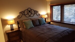 Une chambre à coucher comprenant un lit, deux tables de chevet avec des lampes, un élément décoratif mural et une fenêtre avec des stores.