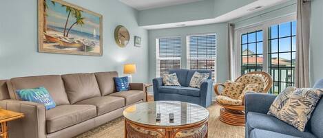 A living room with a beige sofa, a blue armchair, a wicker coffee table, and a painting of a beach scene.