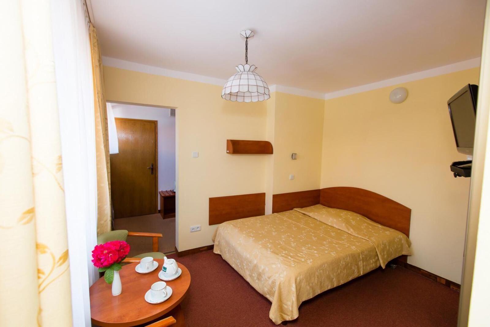 A hotel room with a bed, a small table with a vase of flowers, a bedside table, a wall-mounted TV, and a door leading to another room.