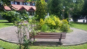 A paved circular area with a wooden bench surrounded by greenery and a building with a red-tiled roof in the background. - Geovita Zlockie (Muszyna)
