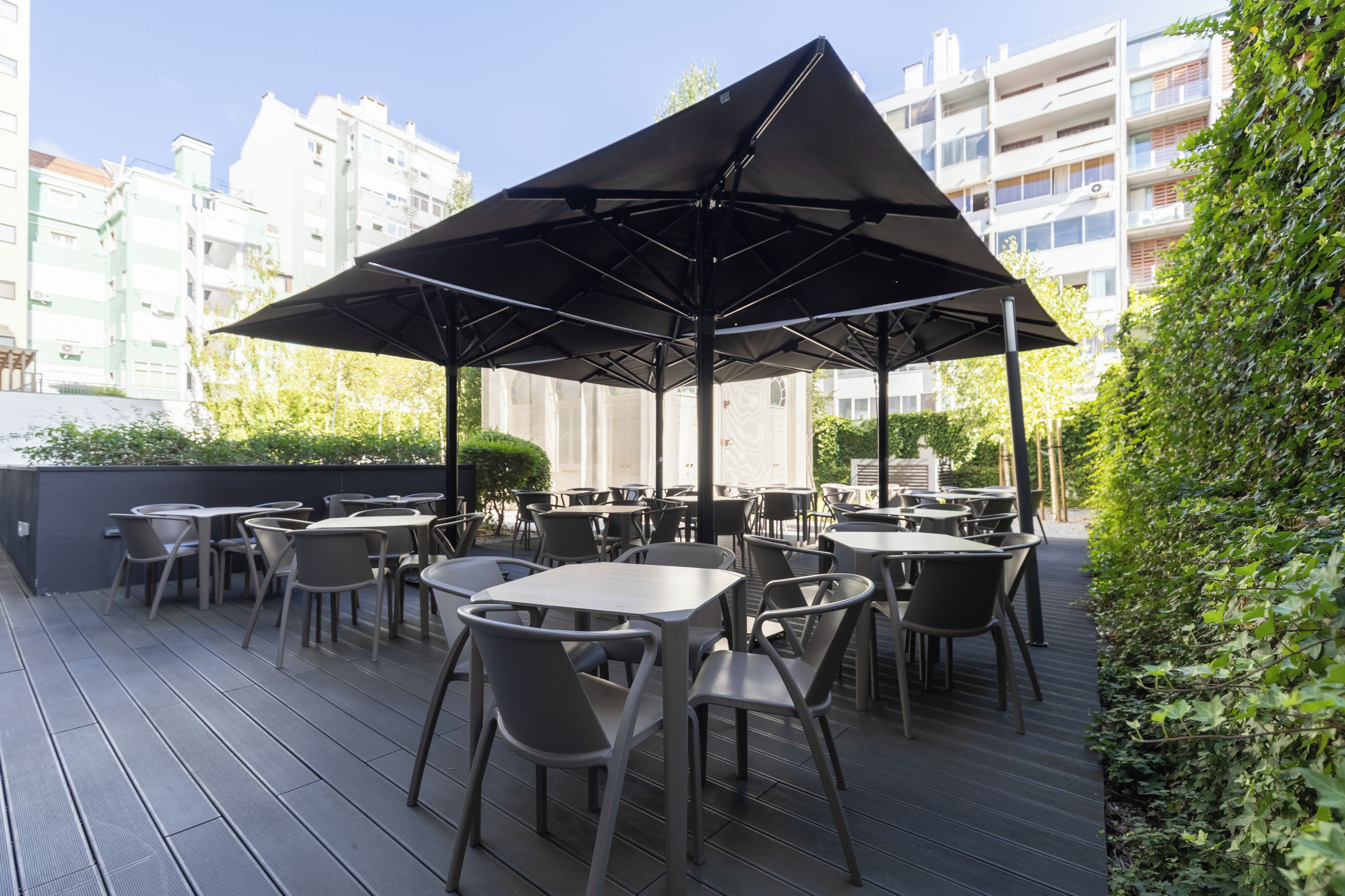 An outdoor seating area with tables and chairs under a large umbrella, surrounded by greenery and buildings.