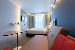 Une chambre d’hôtel moderne équipée d’un lit, d’un bureau, d’un téléviseur à écran plat et d’une grande fenêtre avec des rideaux.