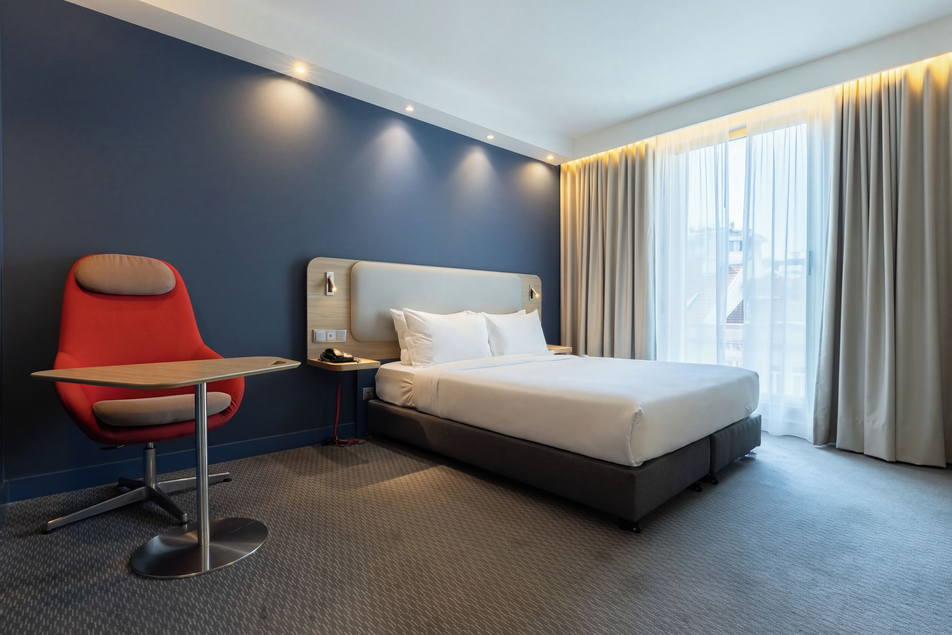 Une chambre d’hôtel avec un lit, un bureau, une chaise et une grande fenêtre avec des rideaux.