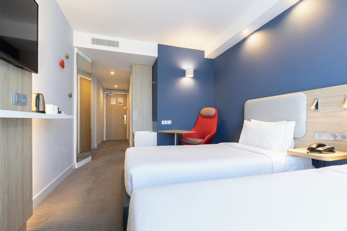 Une chambre d’hôtel avec deux lits, un fauteuil rouge, une table de chevet avec un téléphone et un téléviseur fixé au mur.