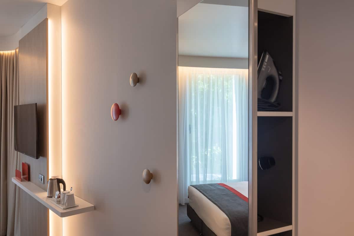 Habitación de hotel con cama, televisión montada en la pared, estante con tetera y tazas, y ventana con cortinas.